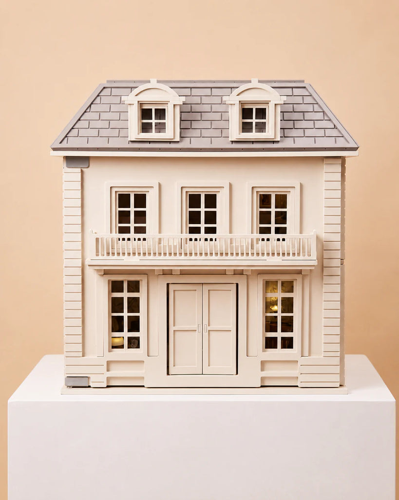 The House of Hamptons — Little Forest Animals, a beige and gray two-story dollhouse featuring a balcony and four dormer windows, is displayed on a white pedestal against a plain tan backdrop, capturing the inviting East Coast style ideal for Little Forest Animals to make their home.