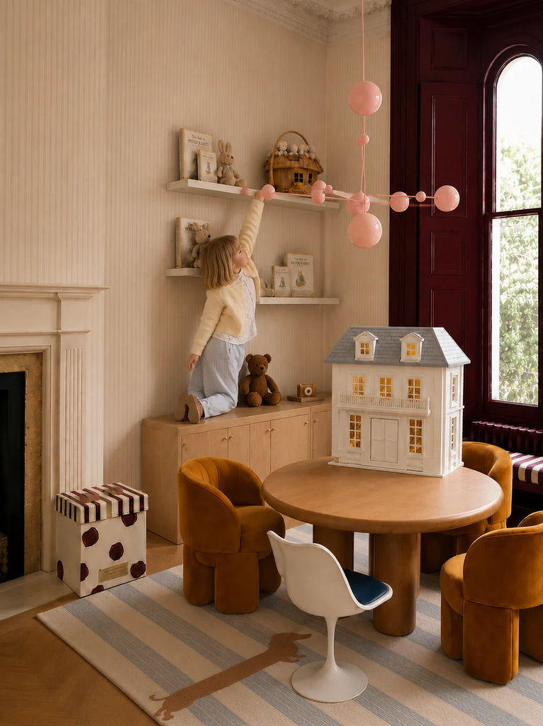 A child stands on a cabinet reaching for a toy on a high shelf in a cozy playroom featuring the House of Hamptons — Little Forest Animals dollhouse, a round table, yellow chairs, and a playful pink chandelier.