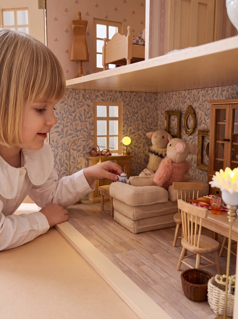 A young child plays with the Fully Furnished Dollhouse, House of Hamptons — Little Forest Animals, arranging miniature furniture and Little Forest Animals in a cozy living room scene complete with a couch, chairs, and soft lighting.