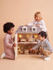 Three young children play together with the House of Hamptons — Little Forest Animals dollhouse, smiling and interacting with its miniature rooms and furniture against a beige background.