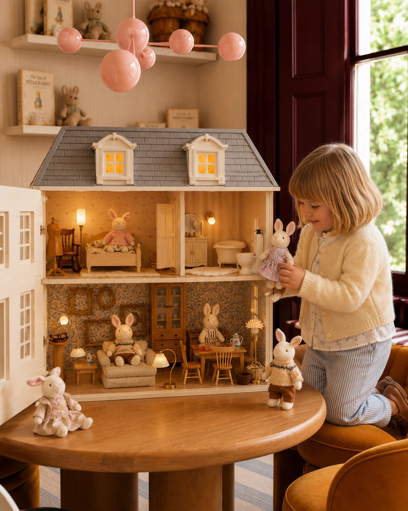 A young child plays with House of Hamptons — Little Forest Animals rabbit dolls in front of a detailed, illuminated East Coast dollhouse adorned with miniature furniture. More Little Forest Animals rabbit dolls from the House of Hamptons collection are inside the dollhouse and on the nearby table.