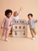 Three young children smile and play around the House of Hamptons — Little Forest Animals, a spacious, detailed dollhouse with glowing lights, set against a beige background.
