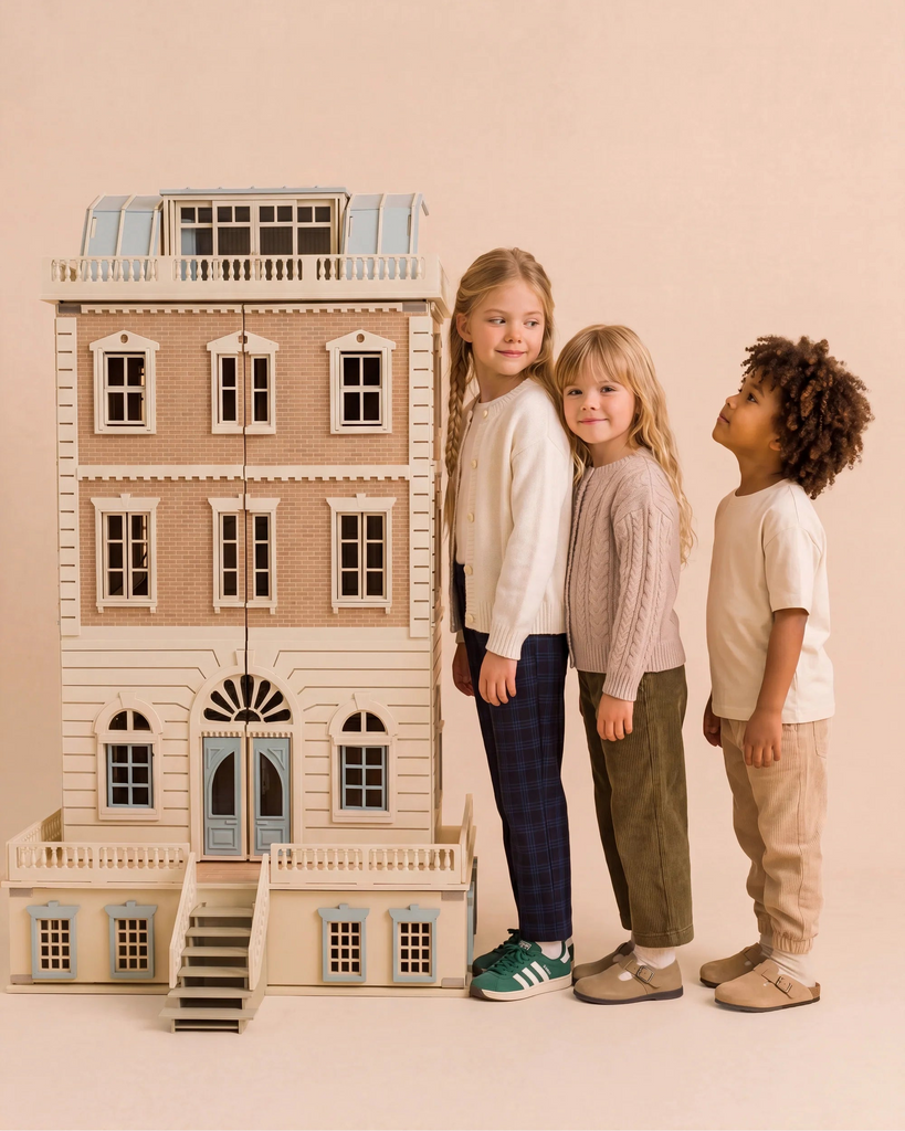 Three children stand in a row beside the House of New York - The Six-Storey Giant, a fully furnished dollhouse with lights. Smiling, two girls are at the front and a boy stands behind them, all dressed in casual, neutral-colored clothing.