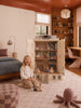 A young child sits and smiles in a cozy bedroom beside the Fully Furnished Dollhouse With Lights, House of New York - The Six-Storey Giant, complete with toy figures. The room features soft colors, a bed, bookshelves, and a plush octopus on the floor.