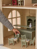 A hand arranges miniature green chairs and a table with tiny dishes inside the fully furnished House of Paris Luxury Dollhouse – A Tale of Parisian Magic, featuring large windows and checked flooring.