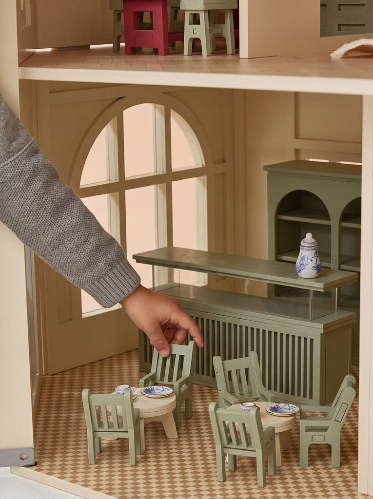 A hand arranges miniature green chairs and a table with tiny dishes inside the fully furnished House of Paris Luxury Dollhouse – A Tale of Parisian Magic, featuring large windows and checked flooring.