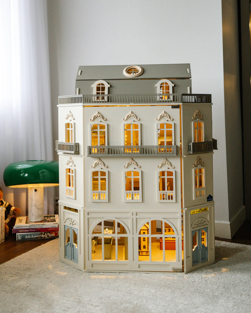 The House of Paris Luxury Dollhouse – A Tale of Parisian Magic stands in a cozy room, its lit windows casting a warm glow as books and a green lamp sit nearby.