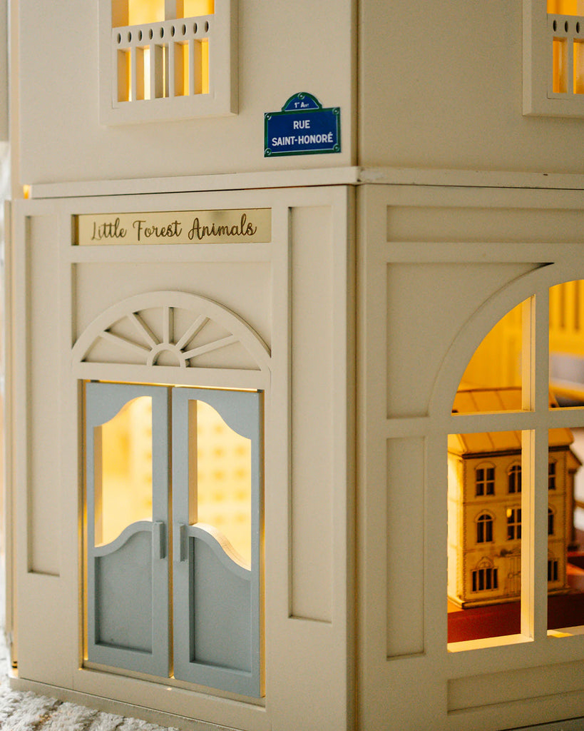 The House of Paris Luxury Dollhouse – A Tale of Parisian Magic features a cream-colored, fully furnished Parisian design with a blue "Rue Saint-Honoré" street sign, an arched doorway labeled "Little Forest Animals," and visible miniature interiors beautifully illuminated from within.