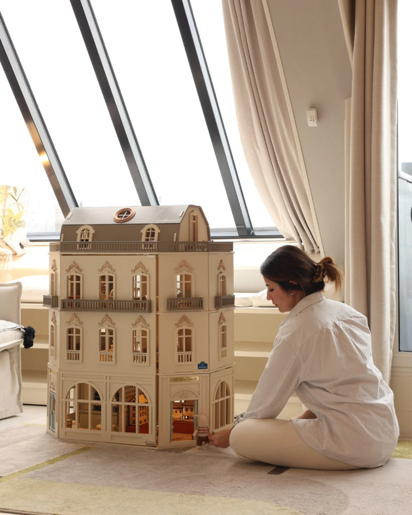 A woman dressed in light clothing sits on the floor beside the House of Paris Luxury Dollhouse – A Tale of Parisian Magic, gently interacting with its door. She is indoors, next to a window with slanted glass panels and beige curtains.