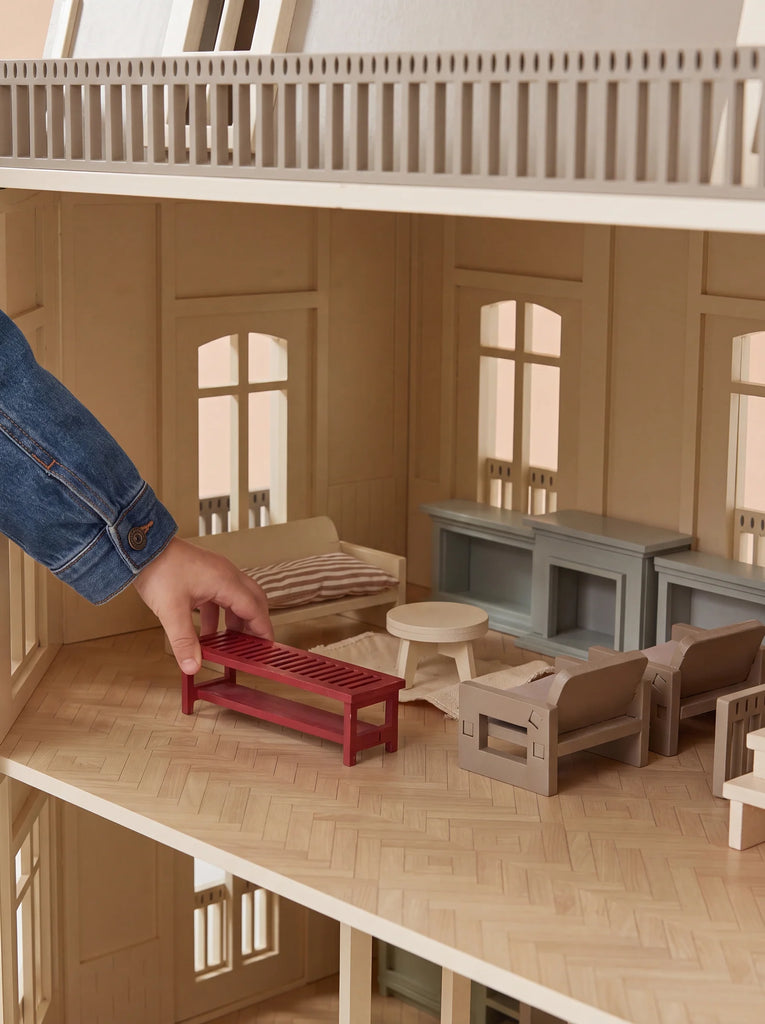 A person's hand in a denim sleeve places a small red bench inside the fully furnished living room of the House of Paris Luxury Dollhouse – A Tale of Parisian Magic, surrounded by miniature furniture including chairs, a table, and cabinets.