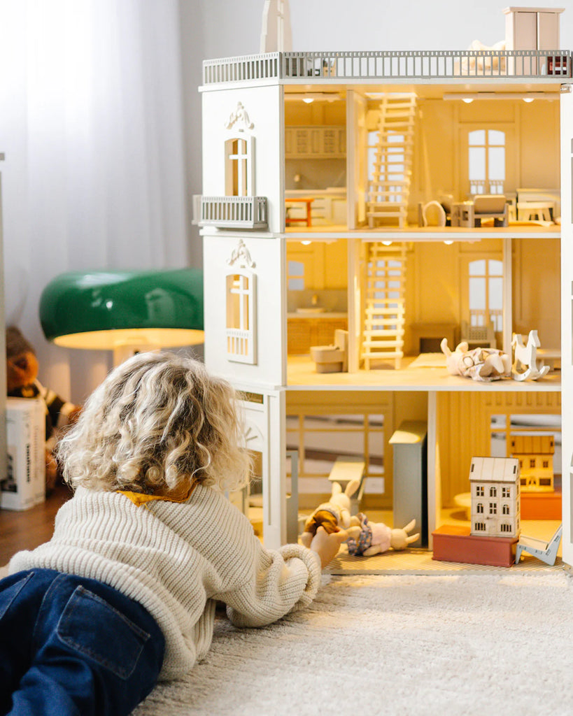 A child with curly blond hair lies on the floor, playing with dolls in front of the House of Paris Luxury Dollhouse – A Tale of Parisian Magic, a large, fully furnished dollhouse featuring multiple rooms and intricate furniture.