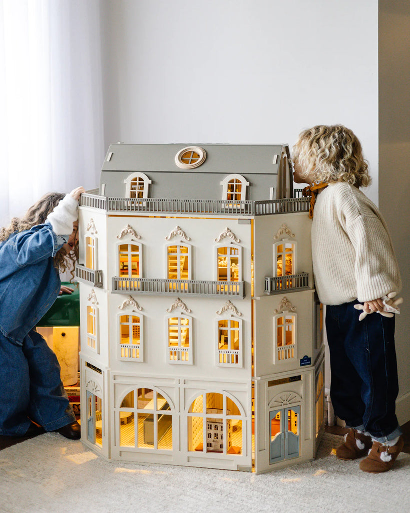 Two children gaze into the House of Paris Luxury Dollhouse – A Tale of Parisian Magic, admiring its spacious, intricately furnished multiple floors and glowing windows. The luxury dollhouse sits on a carpeted floor beside a window draped with white curtains.