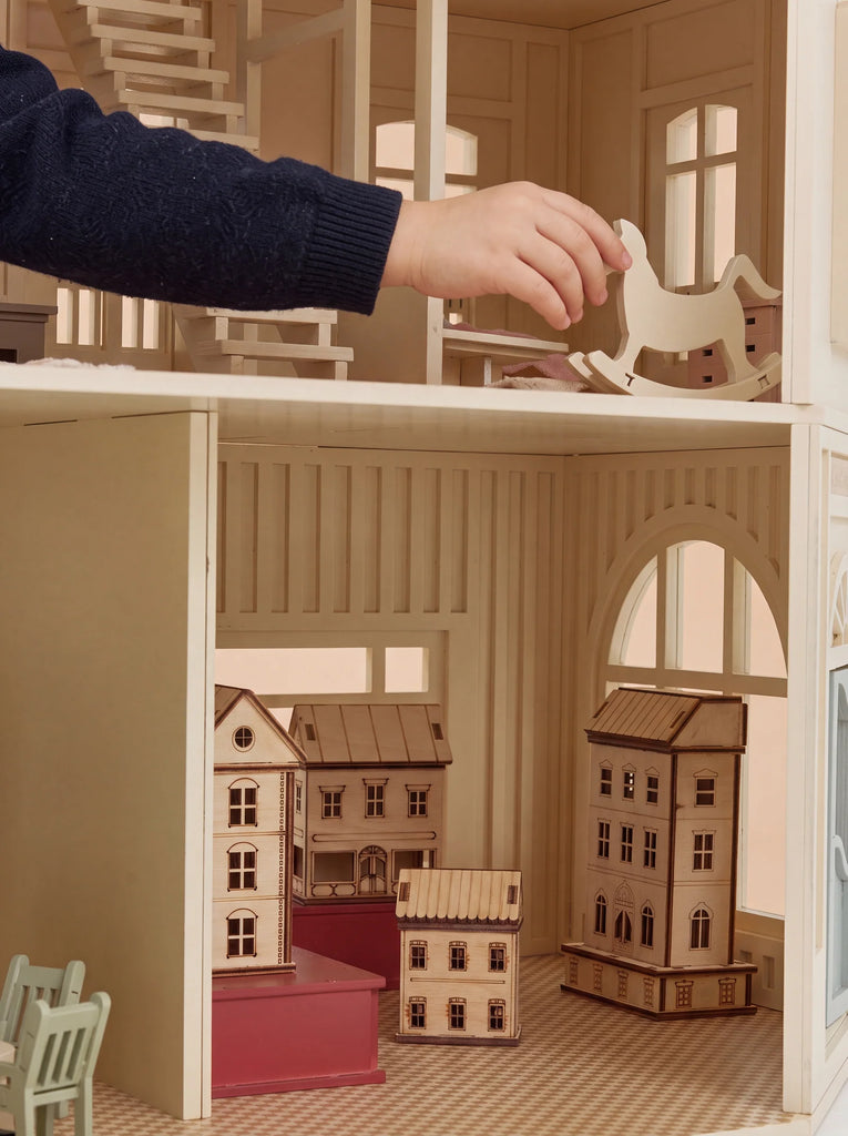 A child’s hand places a wooden rocking horse in the House of Paris Luxury Dollhouse – A Tale of Parisian Magic, where miniature houses and elegantly arranged furniture fill the beautifully furnished rooms.