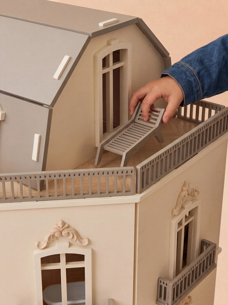 A child’s hand places a miniature lounge chair on the balcony of the House of Paris Luxury Dollhouse – A Tale of Parisian Magic, a detailed, cream-colored dollhouse with gray trim and ornate windows.