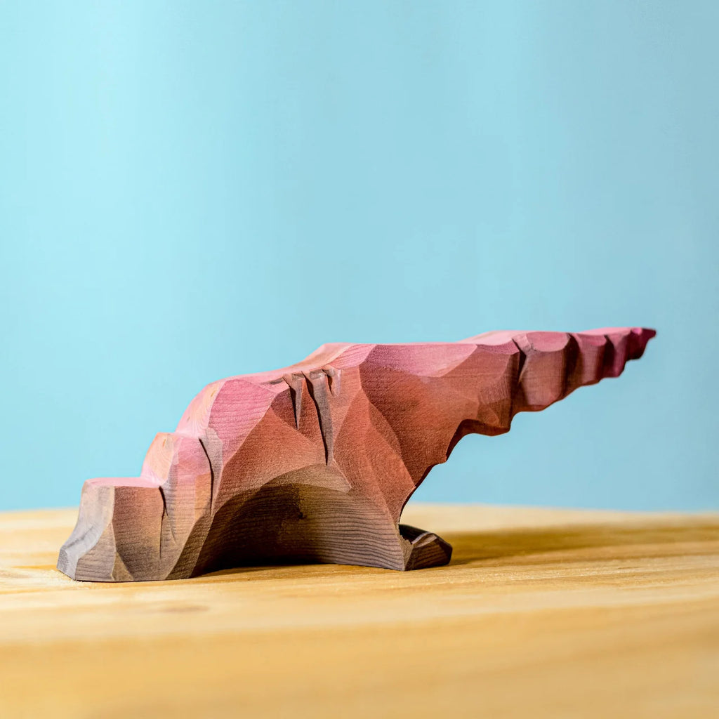 The Bumbu Handmade Wooden Howl Stone—a small, handcrafted wooden figure painted in earthy shades of brown, red, and purple to resemble a rocky cliff or formation—sits on a light wooden surface against a blue background, making it perfect for imaginative play or as a unique wooden toy.