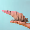 A hand holds the Bumbu Handmade Wooden Howl Stone, a jagged abstract sculpture featuring handcrafted shades of pink, brown, and gray against a light blue background.