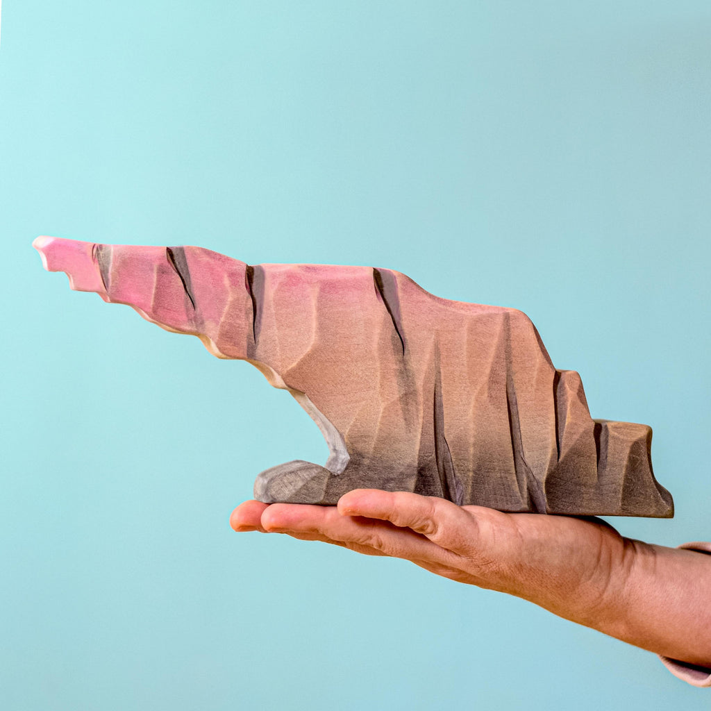 A hand holds the Bumbu Handmade Wooden Howl Stone, a jagged abstract sculpture featuring handcrafted shades of pink, brown, and gray against a light blue background.