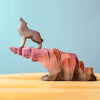The Bumbu Handmade Wooden Howl Stone features a handcrafted wooden sculpture of a howling wolf perched on a jagged, reddish-brown carved cliff, set on a light wooden surface against a plain blue background—perfect for imaginative play or as a unique decorative accent.