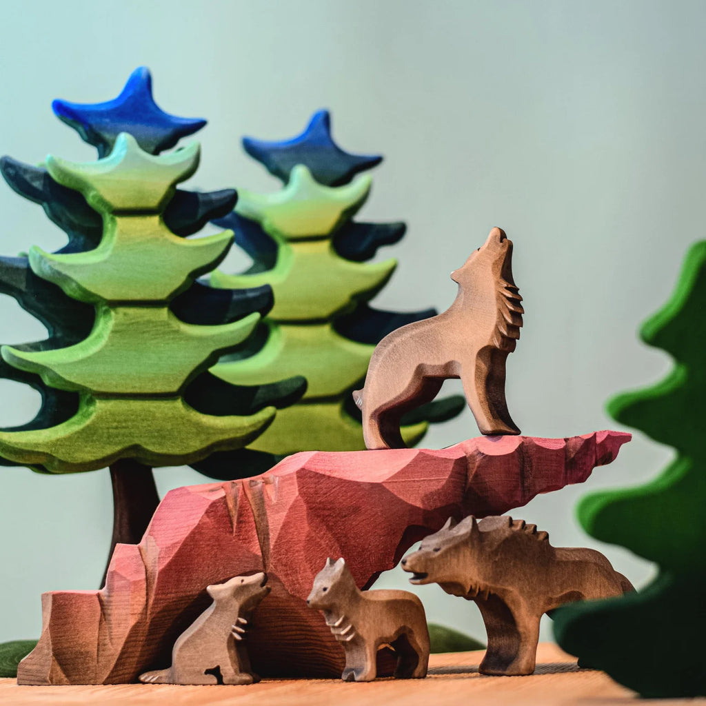 The Bumbu Handmade Wooden Howl Stone features wooden wolf figures positioned on and around a red rock, with three green stylized wooden trees in the background, and a handcrafted wooden toy wolf howling on top—perfect for imaginative play.