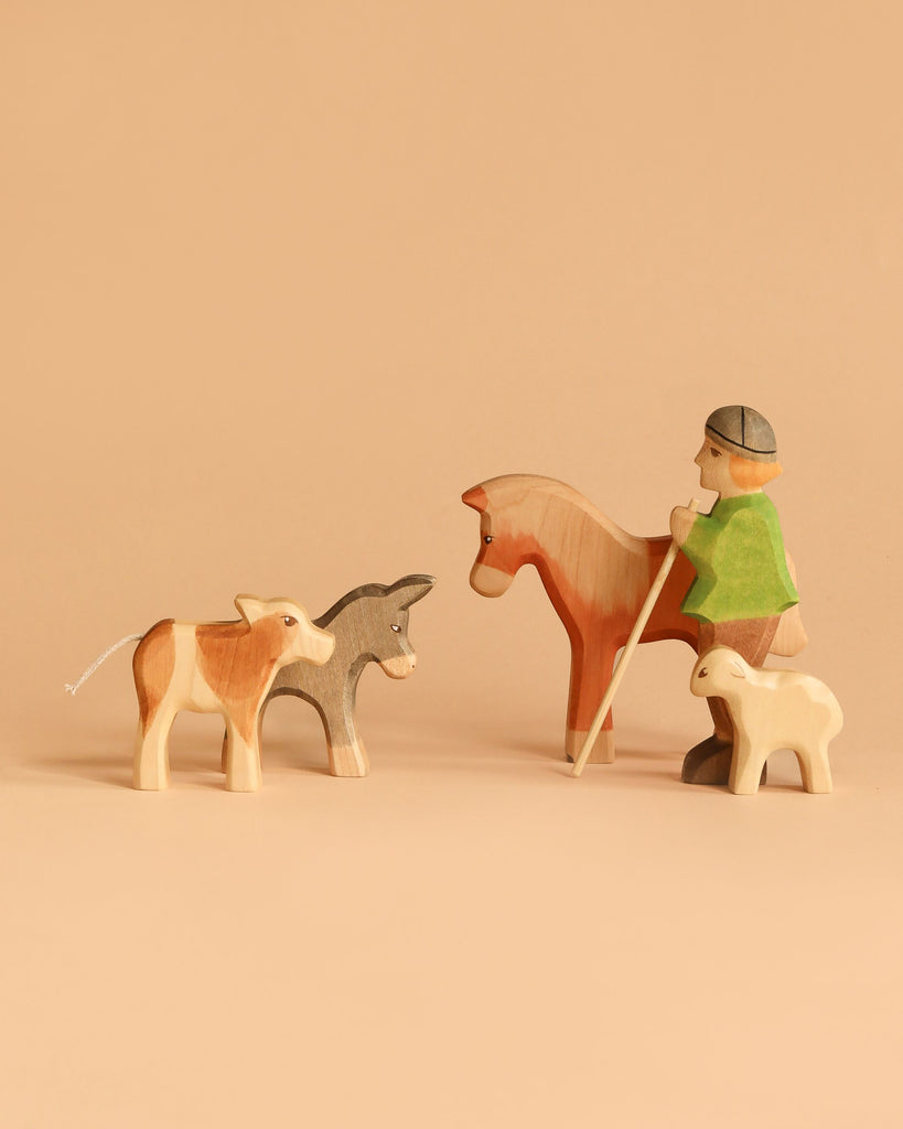 The Handmade Holzwald Shepherd with Farm Animals Set features a shepherd with a staff riding a horse, alongside wooden cow, donkey, and lamb figures, all handcrafted and displayed against a plain beige background.