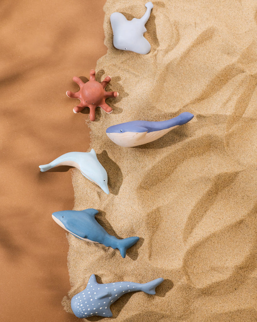 An assortment of colorful ceramic sea creatures including a coral, starfish, whale, and several fish, artfully arranged on a sandy background using non-toxic paint.