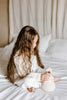 A young girl with long wavy hair sits on a white bed, wearing a cozy white outfit, focused on a Handmade Musical Roly Poly - Lamb she holds in her hands, exploring its musical sounds.
