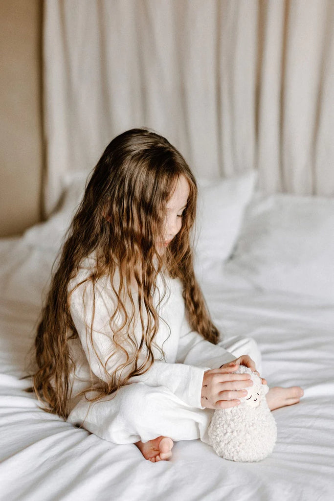 A young girl with long wavy hair sits on a white bed, wearing a cozy white outfit, focused on a Handmade Musical Roly Poly - Lamb she holds in her hands, exploring its musical sounds.
