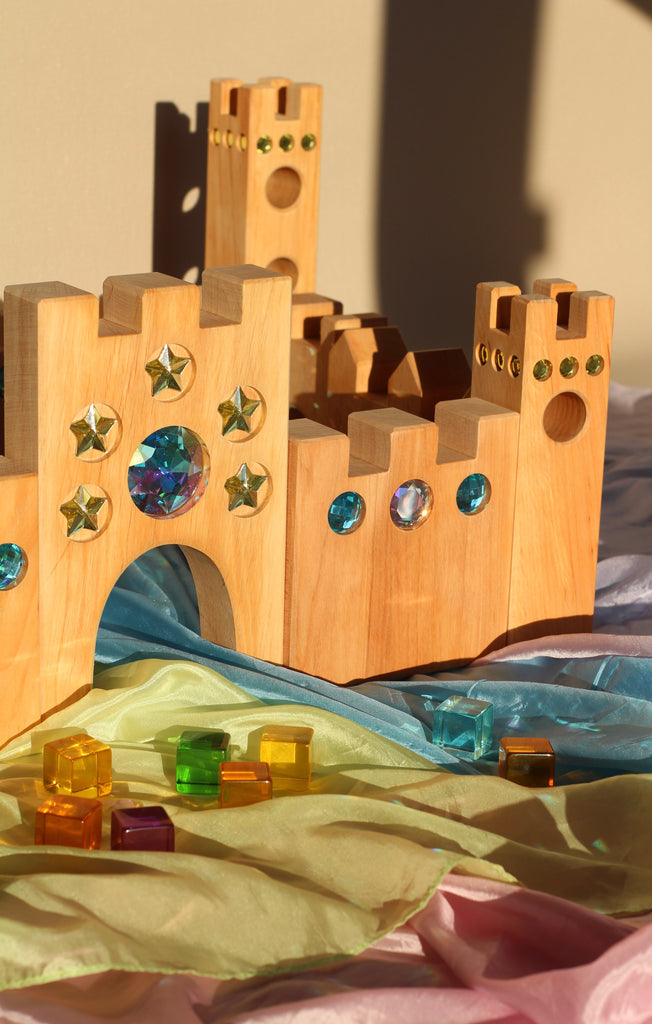 A Bauspiel Large Fairytale Castle, 16-Piece Set made of wood with crenellated towers and inlaid star and circular gems sits on colorful fabric scattered with translucent cubes designed for imaginative play.