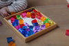 A wooden tray filled with colorful, translucent Bauspiel Lucent Cylinders, 64-Piece Set sits on a table next to a child’s legs.