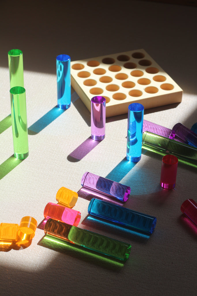 Translucent, colorful Bauspiel Luminous Pillars, 25-Piece Set cylindrical blocks scattered on a surface with a wooden grid base and long, vibrant shadows.