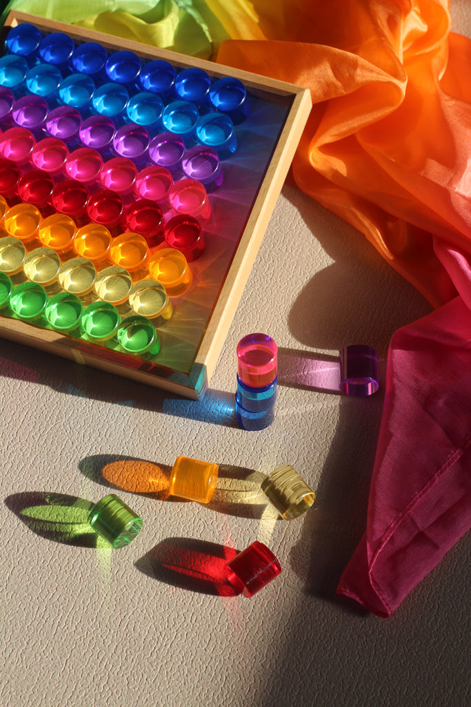 Sixty-four translucent Bauspiel Lucent Cylinders, 64-Piece Set arranged in a wooden tray, with additional pieces scattered nearby casting colorful shadows on the surface next to flowing fabric.