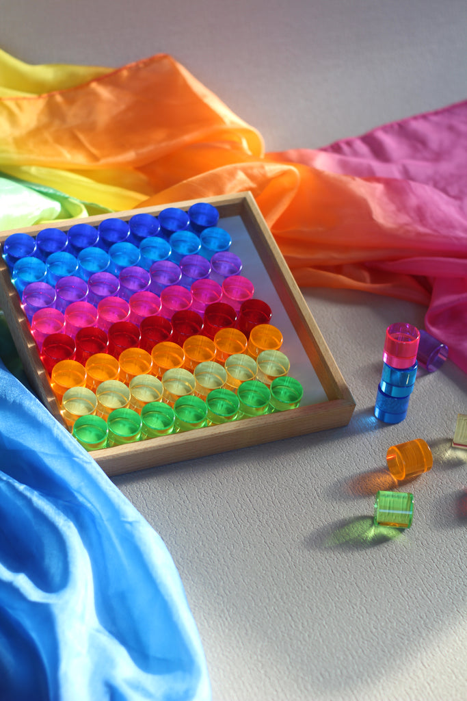 A wooden tray filled with rows of translucent, rainbow-colored Bauspiel Lucent Cylinders, 64-Piece Set, with a stack and a few loose pieces beside it, set against colorful fabric.