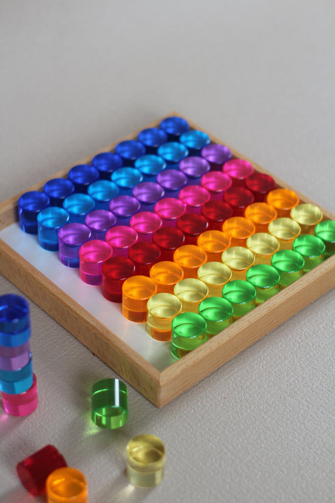 Sixty-four translucent Bauspiel Lucent Cylinders arranged by color in a square wooden tray, with a few extra cylinders standing or lying on the surface nearby.