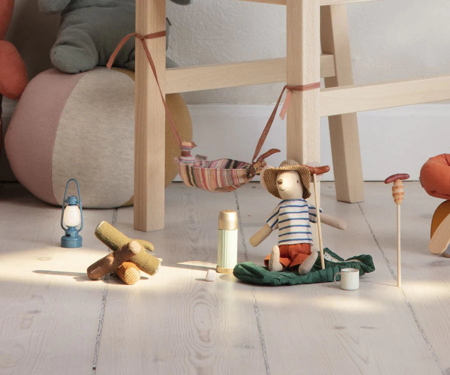 A cozy children's play area with Maileg Campfire, wooden toys and a small lantern on a light wooden floor, next to a low table and a plush cushion.