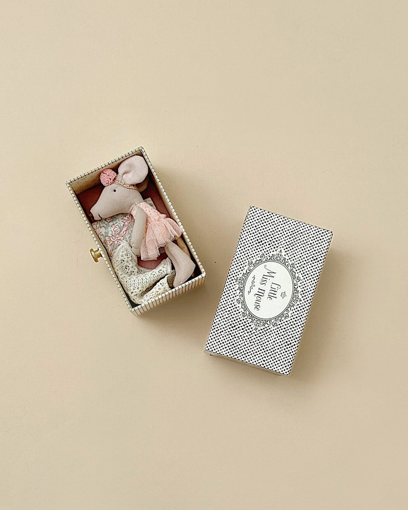 The Maileg Dancing Mouse in Daybed features a little sister mouse lounging in a miniature box decorated with a floral pillow, wearing a pink tutu and headband. A matching polka dot lid rests nearby on the beige surface.