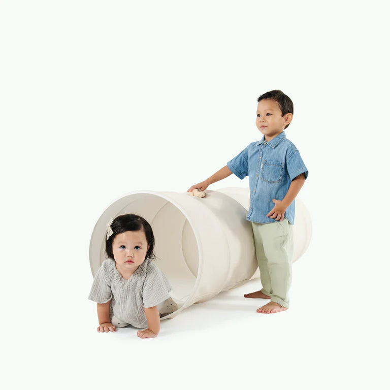 A young boy in a blue shirt and a girl in a gray blouse play with a Gathre Vegan Leather Play Tunnel, on a plain white background. The girl is peeking out from the tunnel while the