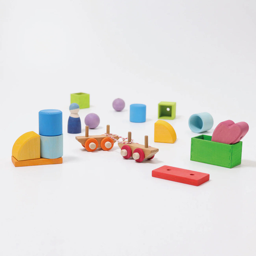 Grimm's Wooden Train Building Set is playfully scattered on a white background, with colorful blocks, peg dolls, carts, and loose parts. Some pieces are stacked near a green box—perfect for inspiring imaginative travel adventures.