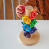 A child's hand places a red ring onto the Grimm's Sorting Helper for Building Rings - Rainbow, a wooden stacking toy with colorful rings on a central rod atop a white table.