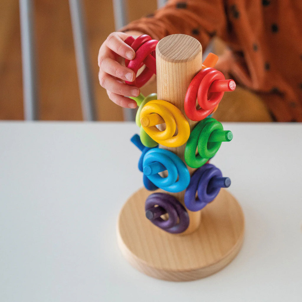A child's hand places a red ring onto the Grimm's Sorting Helper for Building Rings - Rainbow, a wooden stacking toy with colorful rings on a central rod atop a white table.