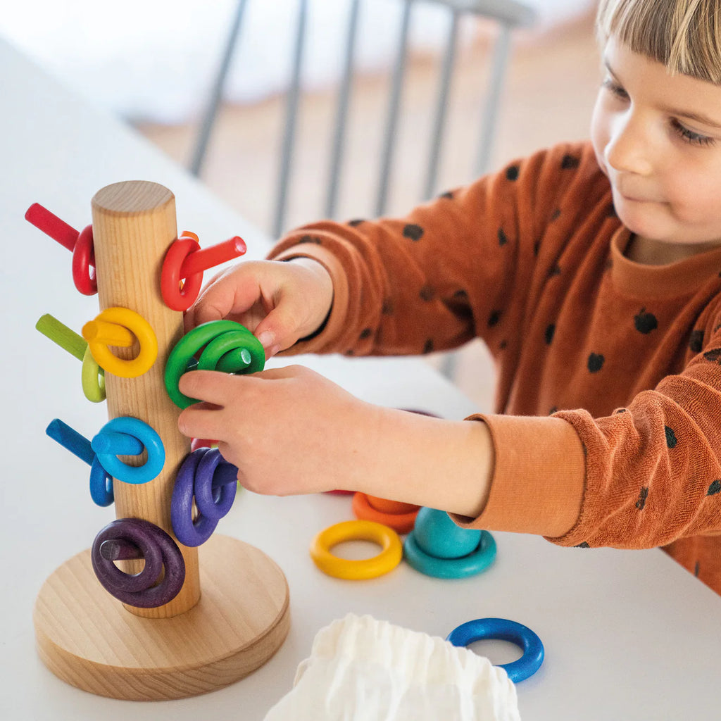 A young child in a brown shirt plays with Grimm's Sorting Helper for Building Rings - Rainbow, stacking colorful rings onto pegs on a vertical stand at a white table.