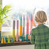 A young child in a green plaid shirt stacks Grimm's Large Building Rollers - Pastel (coming soon) by the window, sunlight shining in and a green plant nearby, building motor skills through this colorful stacking game.