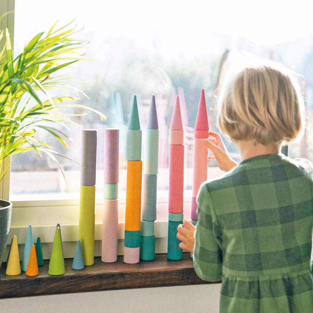 A young child in a green plaid shirt stacks Grimm's Large Building Rollers - Pastel (coming soon) by the window, sunlight shining in and a green plant nearby, building motor skills through this colorful stacking game.