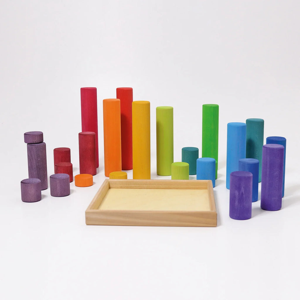 Grimm’s Large Building Rollers Rainbow featuring a wooden tray surrounded by a variety of multicolored cylindrical wooden blocks in different heights and shades, ideal for color sorting and motor skills development.