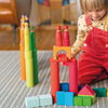 A child uses colorful Grimm’s Large Building Rollers Rainbow and peg dolls to play on a patterned rug, engaging in color sorting.