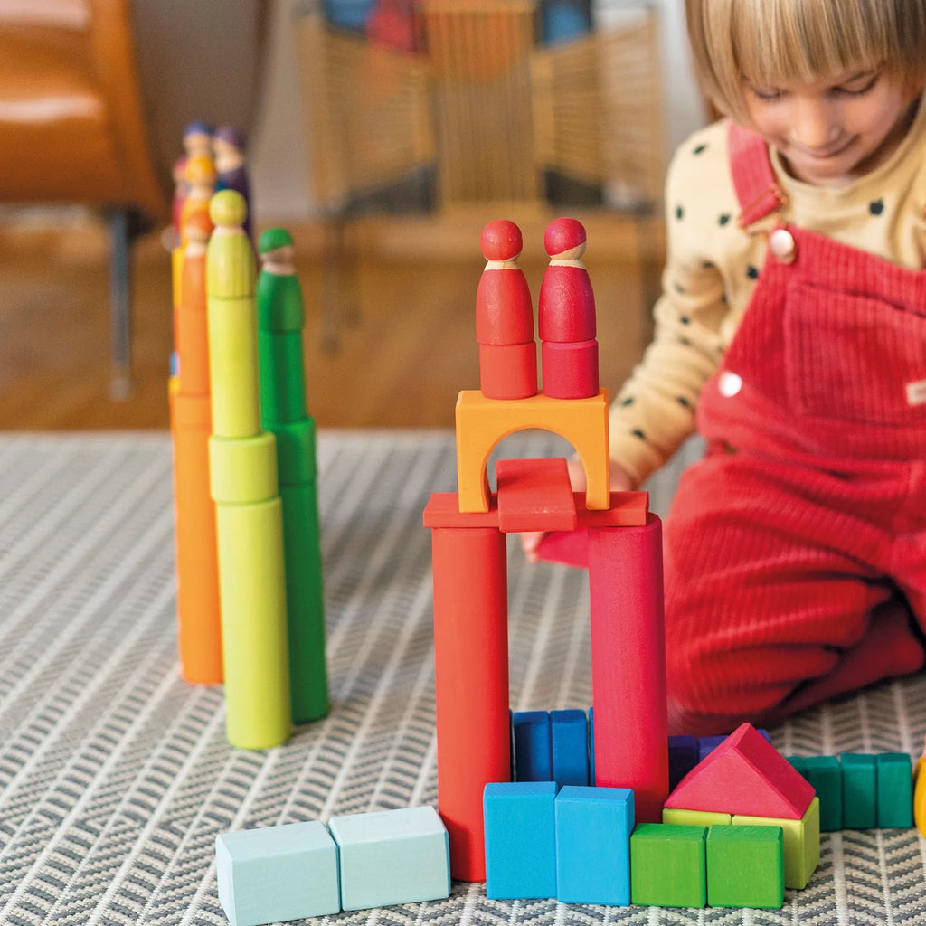 A child uses colorful Grimm’s Large Building Rollers Rainbow and peg dolls to play on a patterned rug, engaging in color sorting.