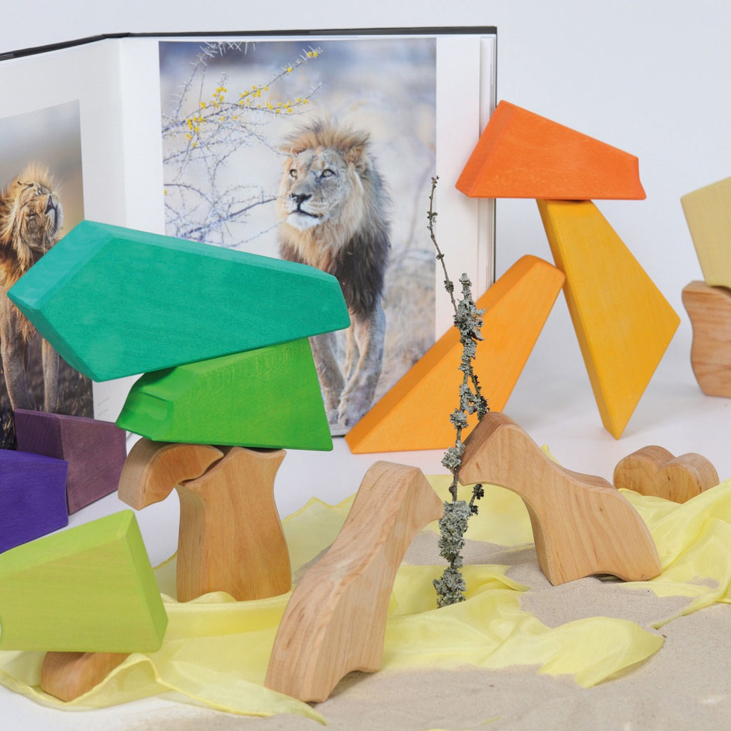 A book featuring a photograph of a lion is open in the background. In the foreground, colorful geometric shapes from Grimm's Rainbow Lion Building Set and natural wood animal figures are arranged creatively. A small branch with lichen is also in the arrangement, with a yellow cloth draped underneath.