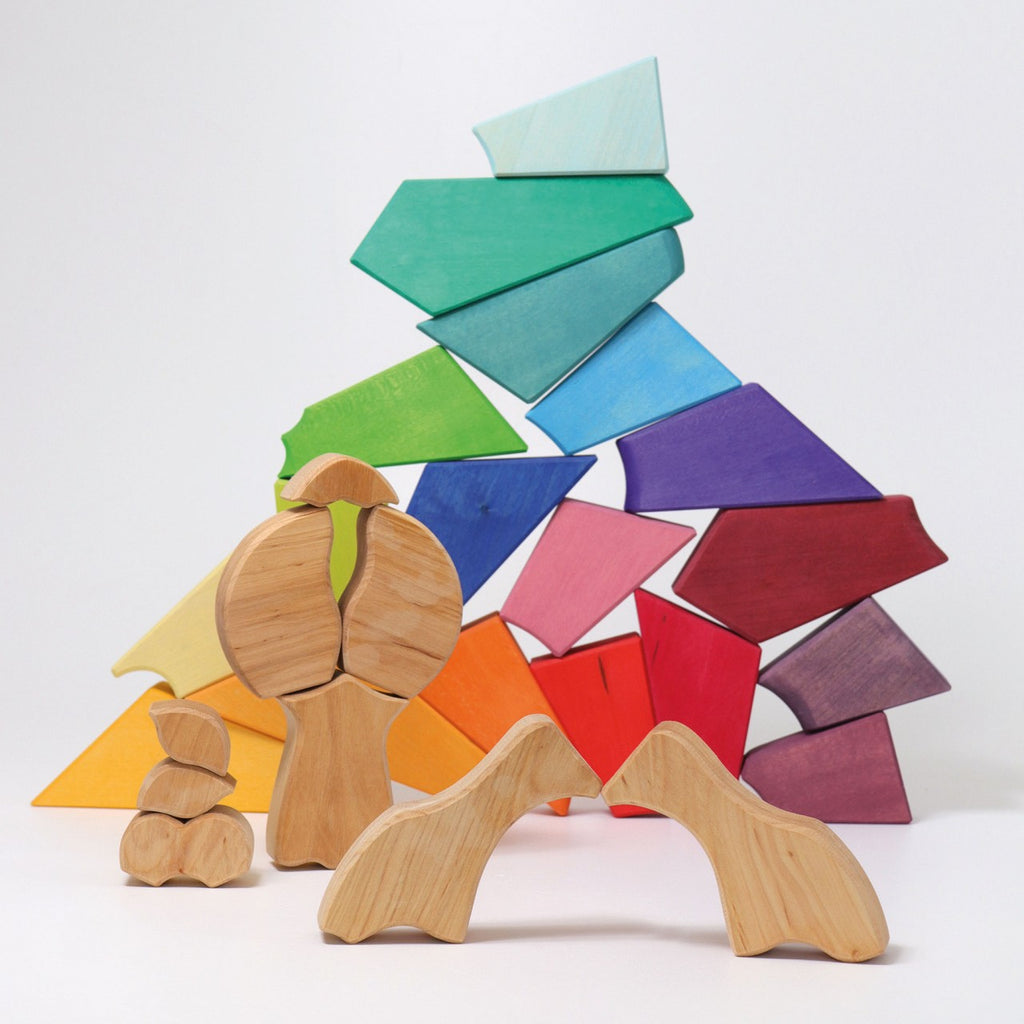 A colorful arrangement of wooden toys forming a creative structure. The vibrant Grimm's Rainbow Lion Building Set includes pieces in various colors like green, blue, purple, red, orange, and yellow. In the foreground are natural wood figures resembling abstract human forms and geometric shapes.
