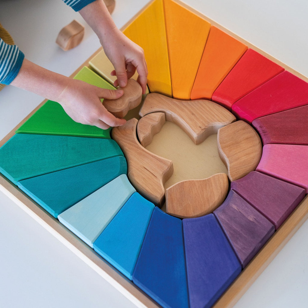 Close-up of a child's hands playing with a Grimm's Rainbow Lion Building Set. The vibrant circular pattern, composed of various colorful geometric shapes in shades of blue, green, yellow, orange, red, and purple, features natural wood pieces and a wooden animal shape in the center.