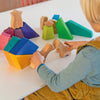 A child is sitting at a white table, playing with a Grimm's Rainbow Lion Building Set. The child is wearing a striped blue shirt and a yellow apron. The natural wood blocks are in various shapes and colors, including green, blue, pink, and yellow.