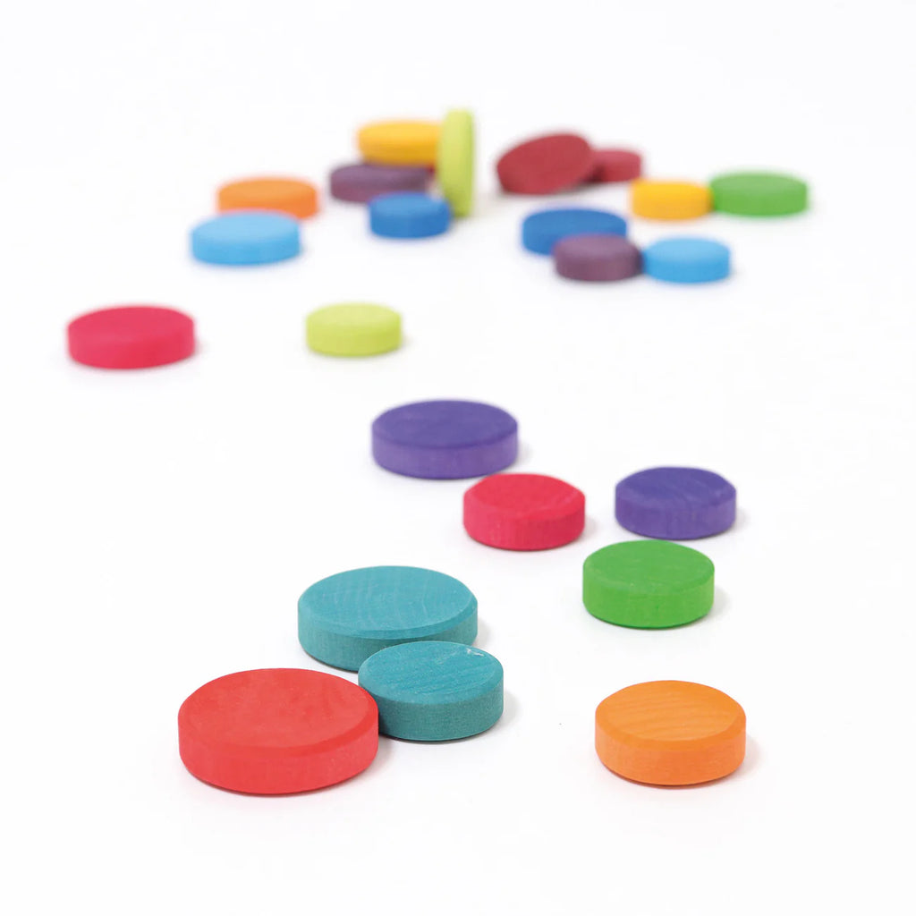 Grimm's Wooden Rainbow Coins (coming soon) are scattered across a white surface, showcasing vibrant red, blue, green, orange, purple, and yellow coins in various sizes near and farther from the viewer.